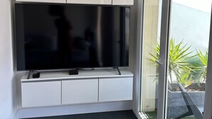Un salon moderne avec un grand téléviseur à écran plat fixé sur un meuble blanc, une porte-fenêtre coulissante donnant sur un balcon agrémenté de plantes et un parquet foncé.