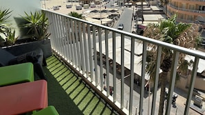 Un balcon avec vue sur une plage, des palmiers et une rue où se trouvent des terrasses de café.