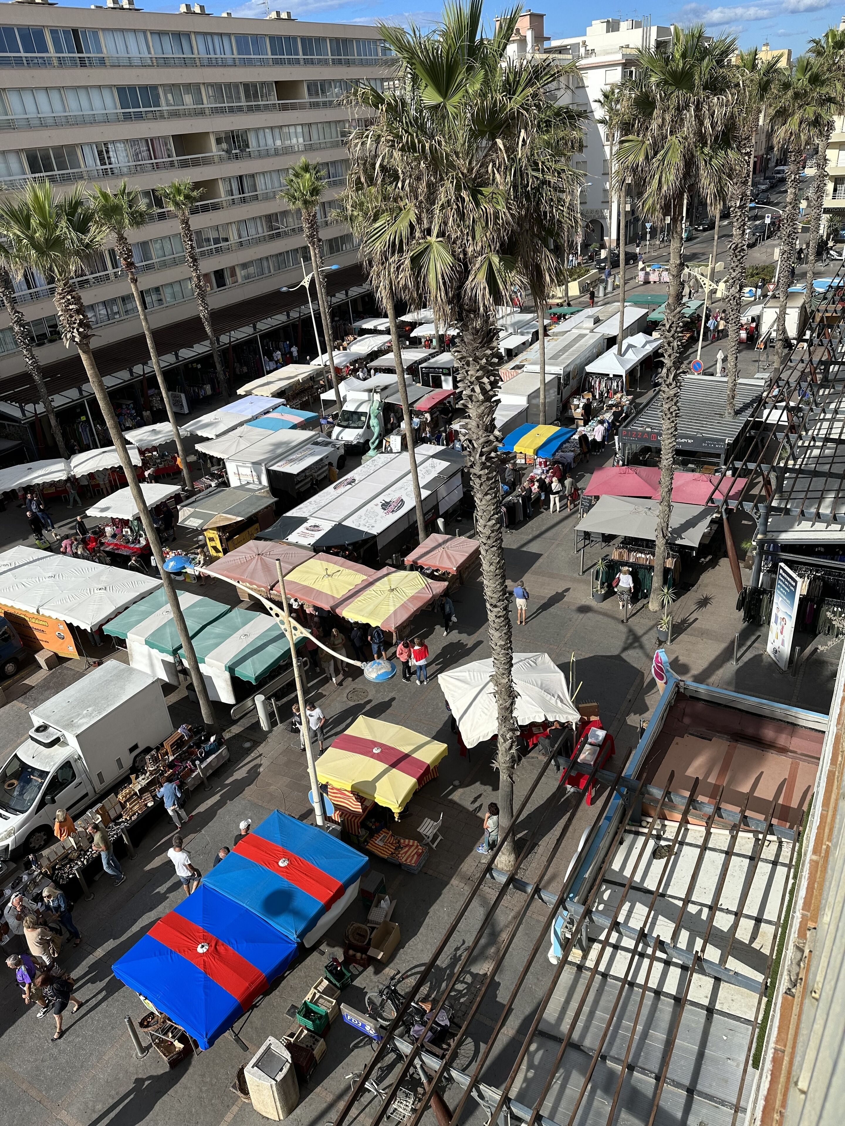 A bustling outdoor market with multiple stalls and vendors, surrounded by palm trees and buildings.