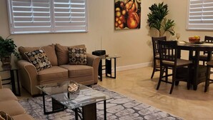 Uma sala de estar com sofá, mesa de centro, mesa de jantar, cadeiras e um quadro na parede.