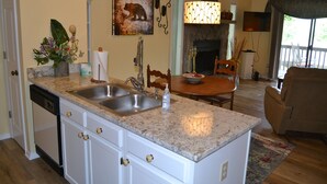 Una cocina con encimera de granito, pileta doble y muebles de cocina blancos. Hay un comedor con una mesa y sillas de madera.