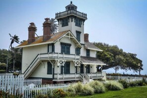 Stately building with a prominent tower, balcony, and staircase, set amidst lush greenery and a white picket fence.