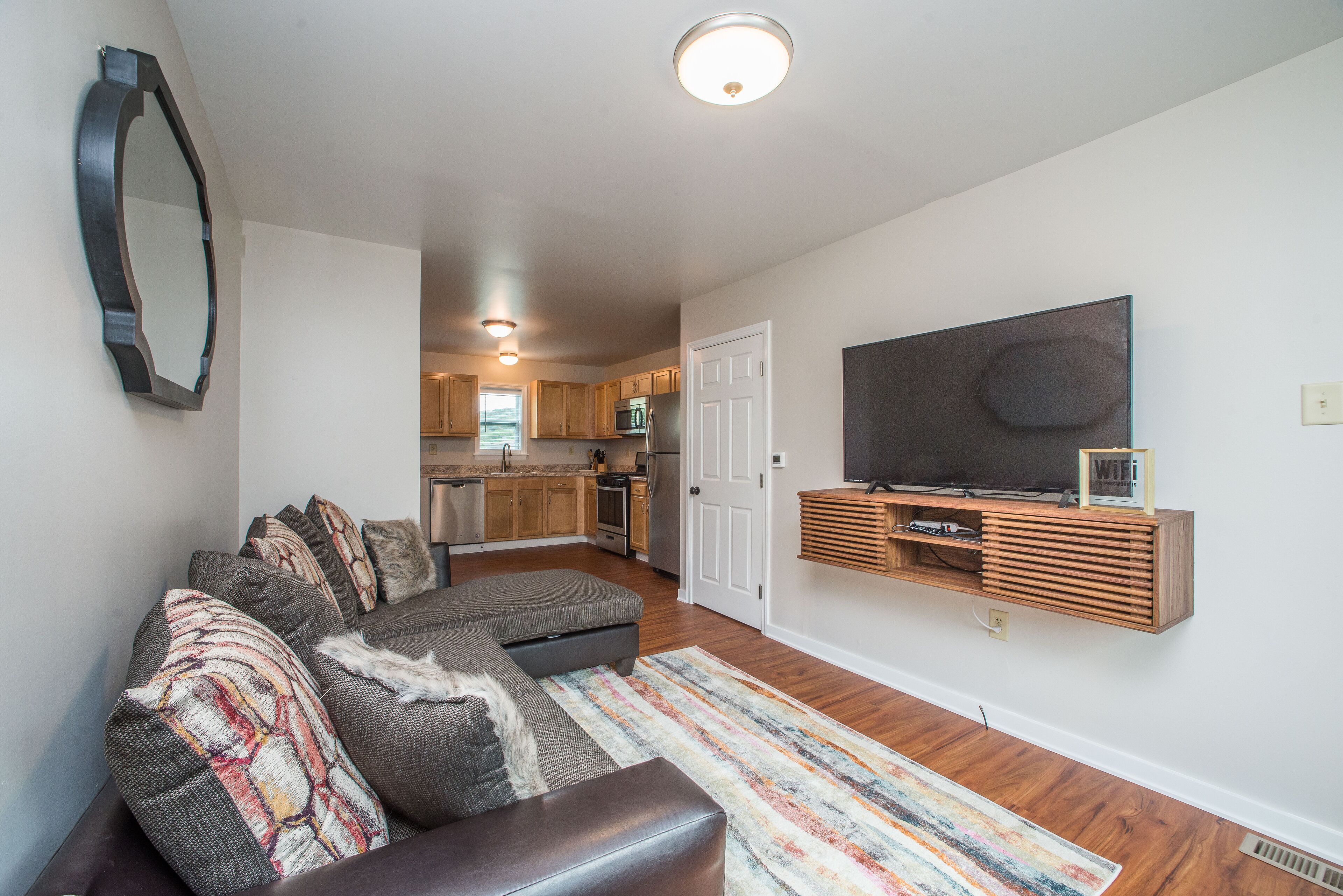 A living room with a sectional sofa, a flat-screen TV on a wooden shelf, and a kitchen in the background.