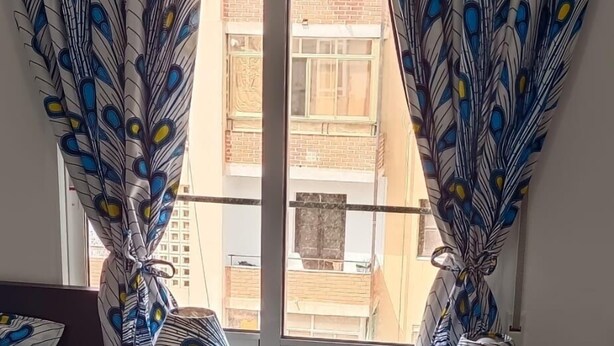 Ein Zimmer mit einem Fenster, das mit pfauenfarbenen Vorhängen verziert ist, einem schwarzen Sofa und einem gemusterten Teppich.