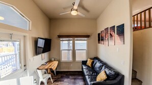 Un espacio habitable compacto con ventilador de techo, televisor montado en la pared, mesa de madera, sofá de cuero negro y ventana con vista a una cerca.