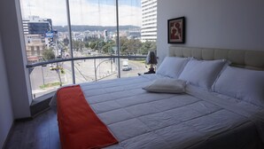 Ein Hotelzimmer mit einem großen Bett, Blick auf die Stadt durch raumhohe Fenster und einem gerahmten Kunstwerk an der Wand.