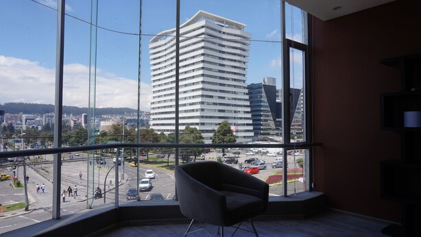 Ein modernes Hotelzimmer mit großen Fenstern und Blick auf die Stadt, darunter ein hohes Gebäude und eine belebte Straße.
