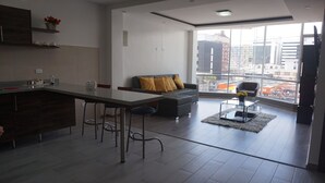 Eine moderne Küche mit Barbereich, ein graues Sofa mit gelben Kissen und ein Blick auf die Stadt durch große Fenster.