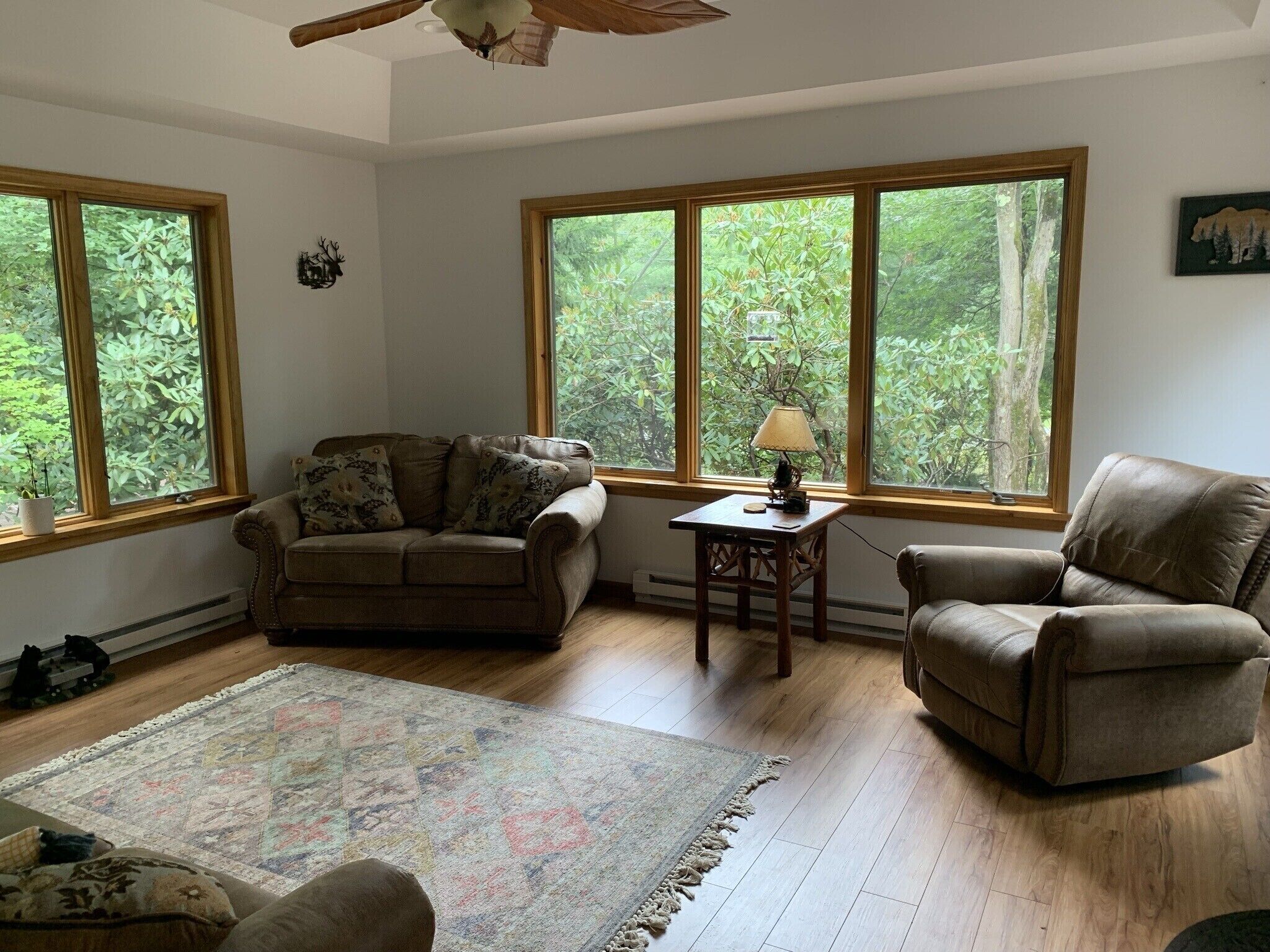 A spacious living room with a large window, a wooden floor, a sofa, a chair, and a small table with a lamp.