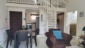 Un vaste espace de vie comprenant un canapé marron, une table à manger et des chaises. Un escalier en colimaçon mène à un niveau supérieur.
