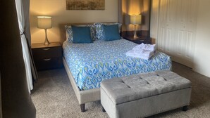 Un dormitorio ordenado con una colcha estampada, dos lámparas de noche, una mesita de noche, un otoman gris y una puerta con cortina.