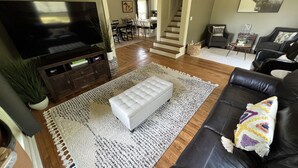 Una sala de estar con televisor, un reposapiés blanco, un sofá de cuero negro y una zona de comedor al fondo.