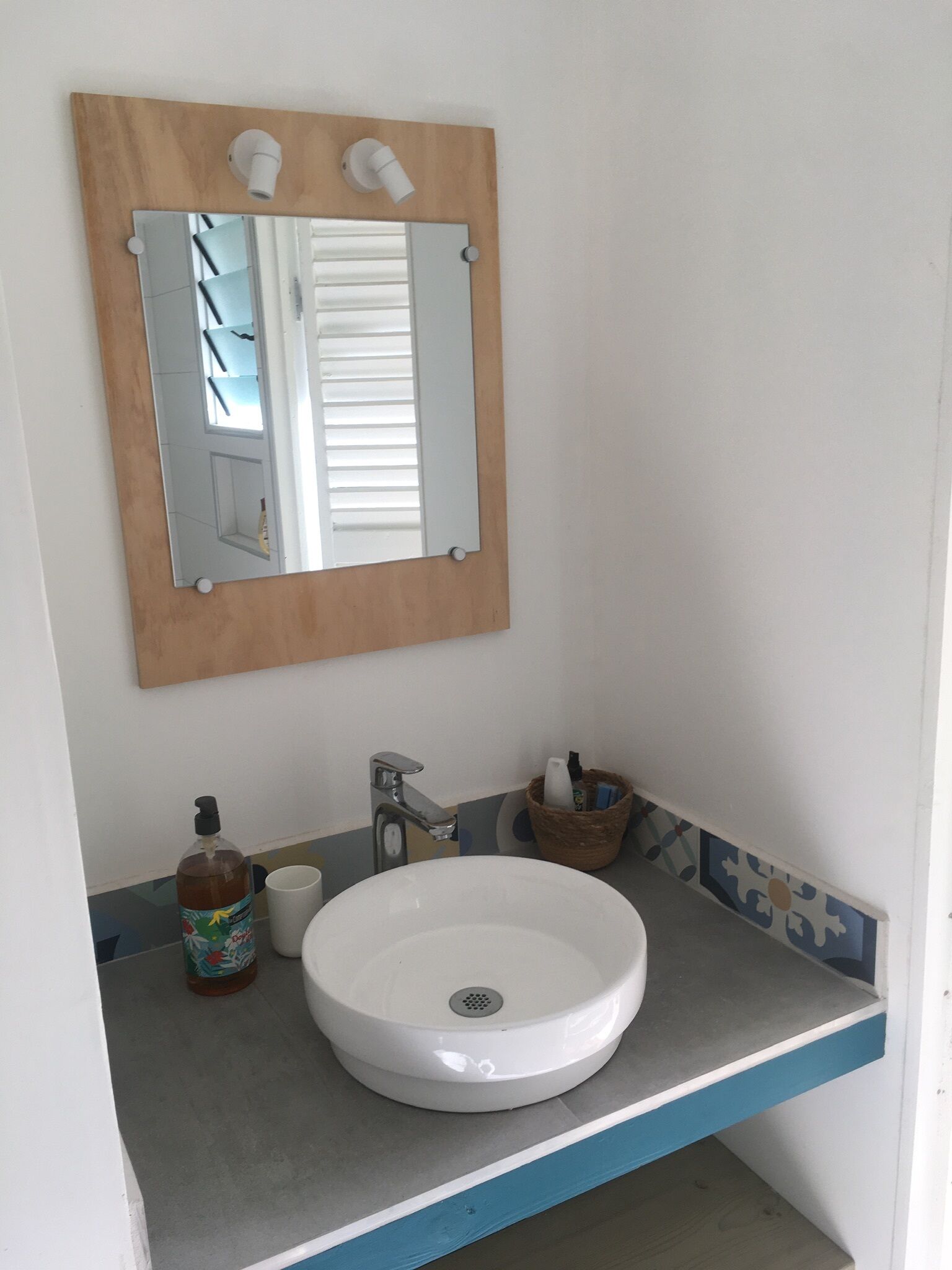 A bathroom with a white sink, a wooden-framed mirror, a faucet, a soap dispenser, and a small basket with toiletries.