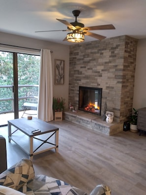 A living room with a fireplace, a ceiling fan, and a view of the outdoors.