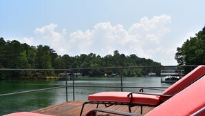 Una terraza de madera con sillones reclinables rojos con vista a un lago, rodeada de árboles y un muelle.
