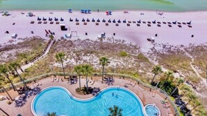 Eine Vogelperspektive zeigt ein Resort mit einem großen, geschwungenen Pool, umgeben von Palmen und Liegestühlen, im Hintergrund ein Strand.