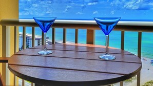 Ein Holztisch mit zwei blauen Martini-Gläsern auf einem Balkon mit Blick auf einen Strand und einen klaren Himmel.