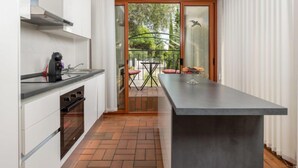 A modern kitchen with a large island, a dining area with a table and chairs, and a sliding glass door leading to a balcony with a view of trees.