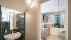 A modern bathroom with a sink, mirror, and dark tiled walls. Adjacent is a kitchen with a microwave, toaster, and wooden cabinets. - WePuglia - Dimore Stefania - Suite Piazzetta (Polignano a Mare)