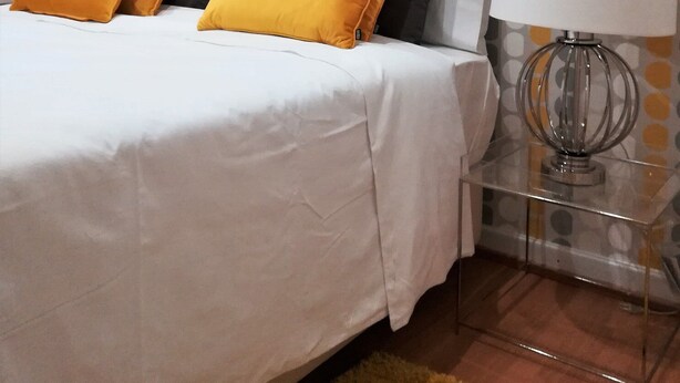 A neatly made bed with white and yellow bedding, a clear glass nightstand with a lamp, and a polka dot wallpaper in grey, white, and yellow. - North Star Flats @dom Luís I Bridge (Vila Nova de Gaia)