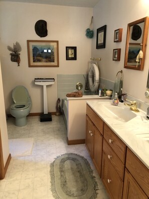 Une salle de bain avec des toilettes, un lavabo et une baignoire. Les murs sont ornés de tableaux encadrés, il y a un porte-serviettes et un tapis sur le sol.
