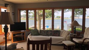 Ein Wohnzimmer mit großem Fenster mit Blick auf ein Gewässer, einem Deckenventilator und einem Fernseher auf einem Holzboden.