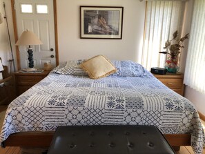 Une chambre avec une couvre-lit à motifs, un tableau encadré au mur, une lampe de chevet et un vase contenant des fleurs séchées.