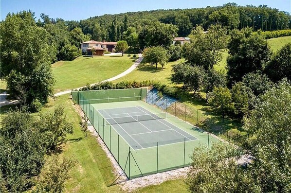 A tennis court surrounded by lush greenery and a few houses in the background.