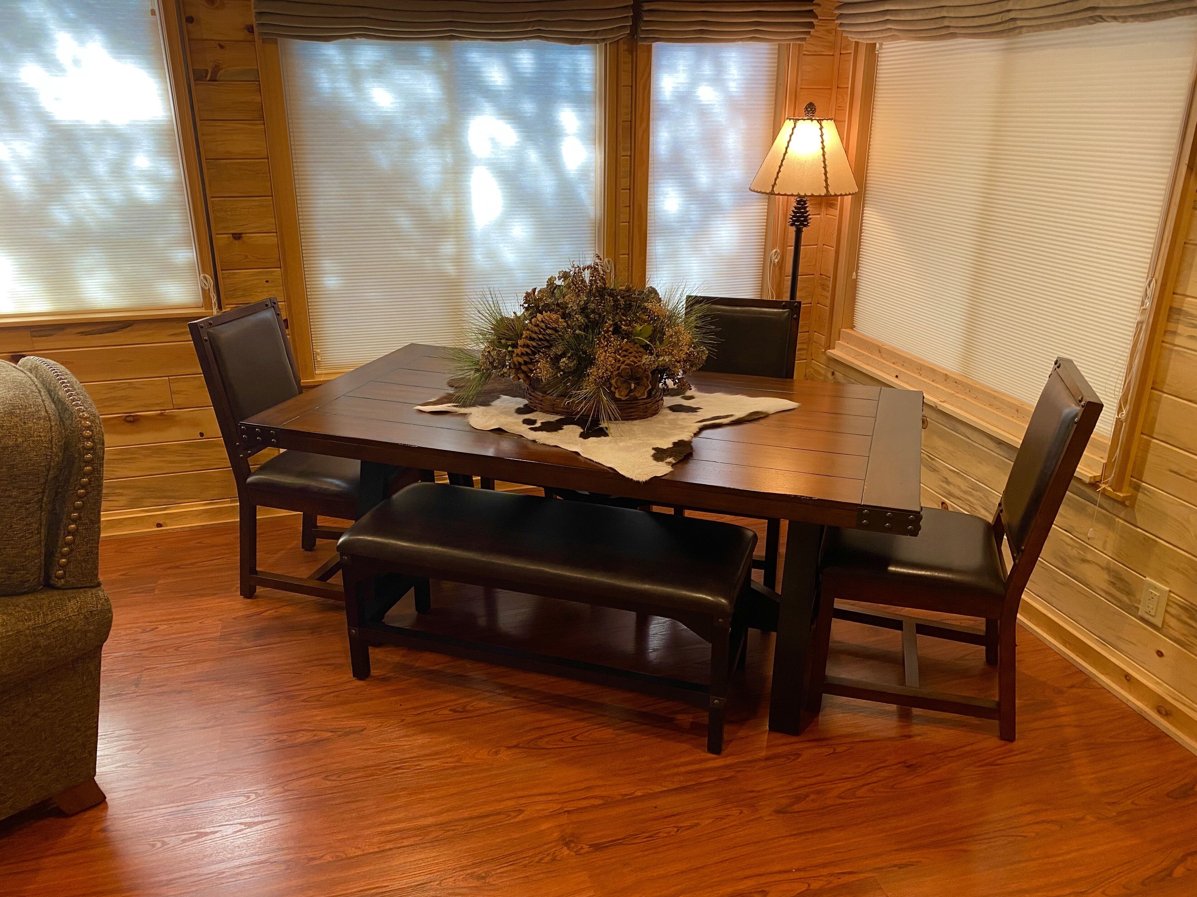 A wooden dining table with a centerpiece, surrounded by chairs, in a room with wooden walls and large windows.