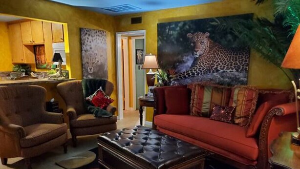 A living room with a red sofa, two armchairs, and a black tufted coffee table. There is a painting of a leopard on the wall and a kitchen area in the background. - Bakersfield Safari Retreat (Bakersfield)