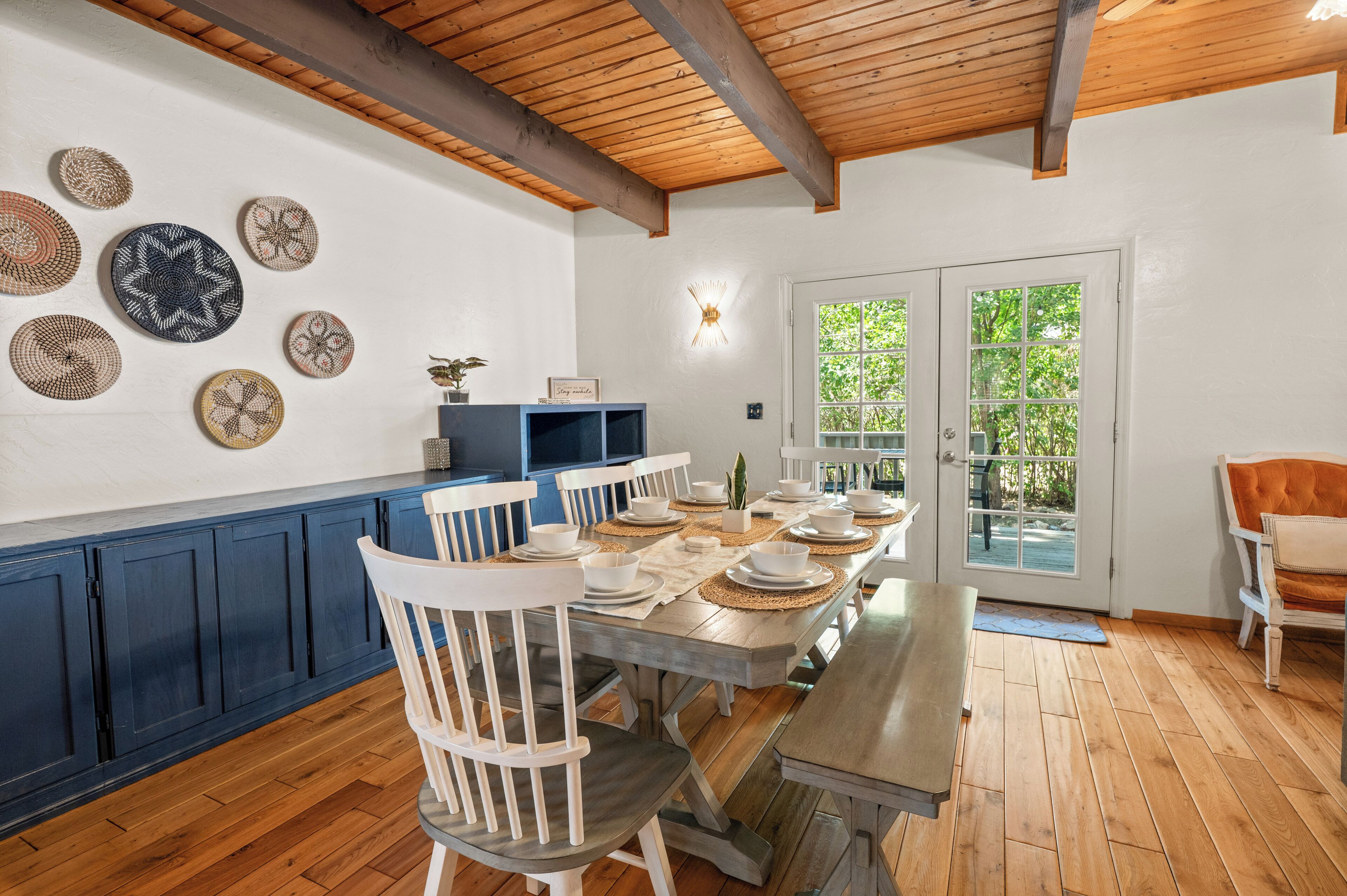 A dining area with a wooden table set for six, surrounded by white chairs. There are decorative plates on the wall and a blue cabinet to the left.