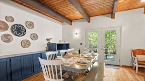 Un coin repas avec une table en bois dressée pour six personnes, entourée de chaises blanches. On peut voir des assiettes décoratives accrochées au mur et un buffet bleu sur le côté gauche.