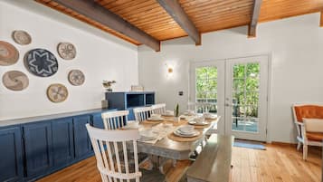 A dining area with a wooden table set for six, surrounded by white chairs. There are decorative plates on the wall and a blue cabinet to the left.