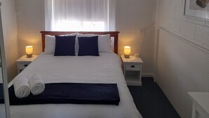 Ein ordentlich bezogenes Bett mit weißer Bettwäsche und zwei dunkelblauen Kissen, flankiert von zwei Nachttischen mit Lampen, ein Fenster mit durchsichtigen Vorhängen und eine hölzerne Fledermaus an der Wand.