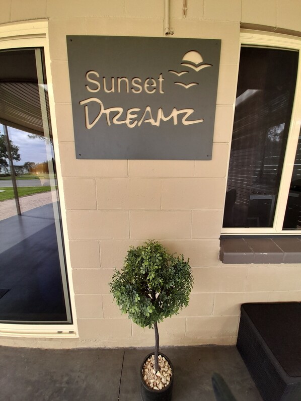 Een bord met de tekst 'Sunset Dreamz' op een beige muur, een kleine pot met een boom en een raam met luiken.