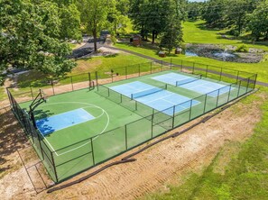 Un court de tennis entouré d’une clôture, avec des arbres et un petit étang à proximité.