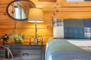 A wooden cabin interior with a bed, a round mirror, a lamp, and a small table with a plant and a vintage coffee pot. - Rustic Log Cabin Near Thunder Ridge w Private Hot Tub & Free Attraction Tickets_Community Amenities! (Ridgedale)