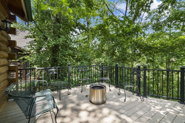 A wooden deck with metal chairs and a metal table, surrounded by a black metal fence, overlooking a wooded area.