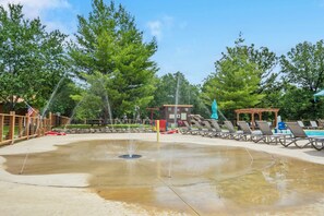 Un espace de jeux aquatiques avec une fontaine, entouré d’arbres et équipé de chaises longues.