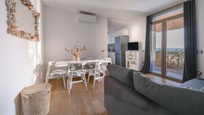 A modern living room with a grey sofa, a dining area with white chairs, and a kitchen in the background. - Casa Frontemare con Scesa al Mare Privata (Castiglioncello (LI))