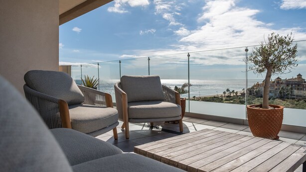 A balcony with two grey upholstered chairs, a wooden table, and a potted plant. - Los Flamencos Penthouse by Ĥ - Haraganes Beach HLHF4501P43 (Isla Canela)