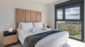 A modern bedroom with a large bed, wooden headboard, and a view of a cityscape through a large window. - Los Flamencos Penthouse by Ĥ - Haraganes Beach HLHF4501P43 (Isla Canela)