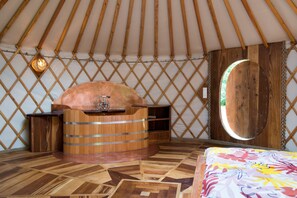 Ein Jurteninneres mit Holzofen, einer Holzbank und einem Bett mit farbenfroher Decke.