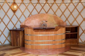 Eine holzgefällte, fassförmige Sauna mit einem Metallhahn, vor einer Spalierwand mit Lichterketten.