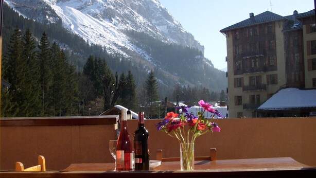 Un balcon avec vue sur des montagnes enneigées, une table en bois ornée d’un vase de fleurs et deux bouteilles de vin.