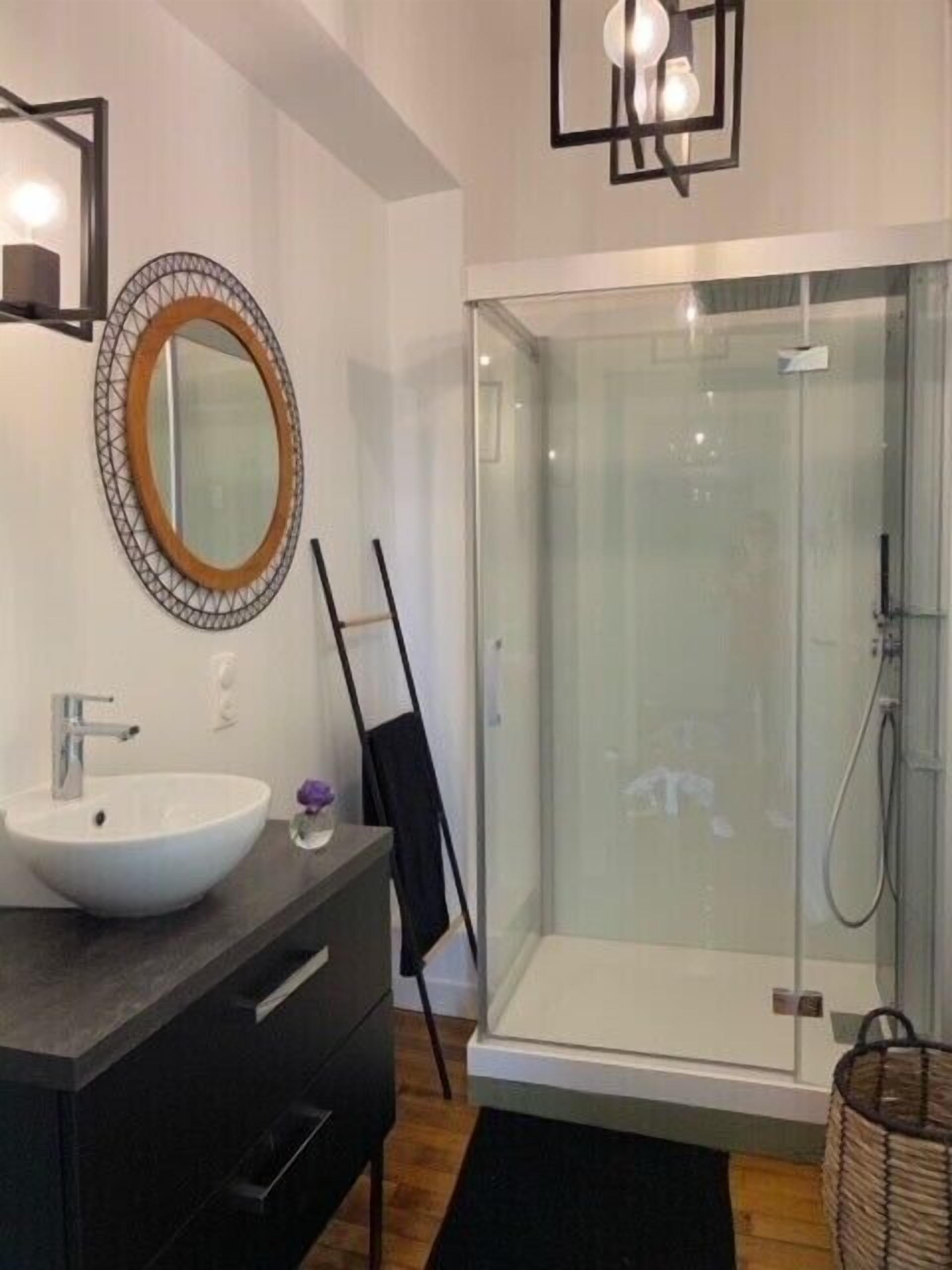 Un baño moderno con un espejo ovalado grande, un lavabo blanco, una silla negra y una ducha con cerramiento de vidrio.