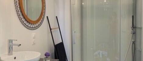 Un baño moderno con un espejo ovalado grande, un lavabo blanco, una silla negra y una ducha con cerramiento de vidrio.