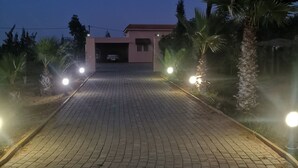 A paved driveway leading to a building with a garage, surrounded by palm trees and illuminated by streetlights. - The Lemon Tree (Ainjohra)