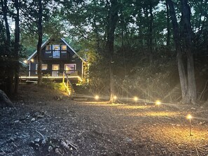 A cabin with a porch, surrounded by trees, and a path with lights leading to it.