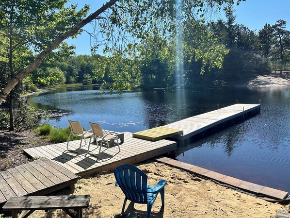 A wooden dock extending into a calm lake, surrounded by trees and a picnic area with chairs and a table.